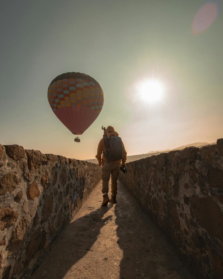 Backpacker walks along an ancient stone path as a hot air balloon drifts in the sky.